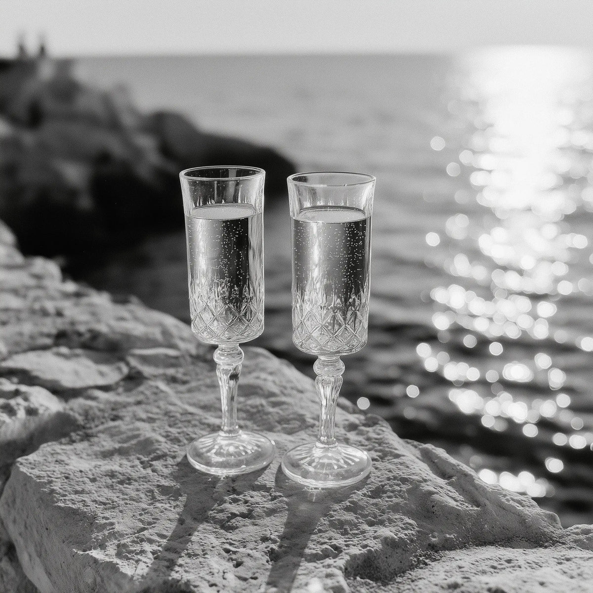 Two clear champagne flutes filled with a bubbly drink, set on a stone ledge with a blurred ocean and rocky shoreline in the background.