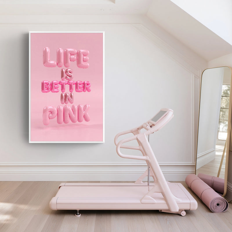 A pink with white frame wall art print with the text 'LIFE IS BETTER IN PINK' hangs on a light wall above a pale pink treadmill in a bright, minimalist room with a mirror and rolled exercise mat.