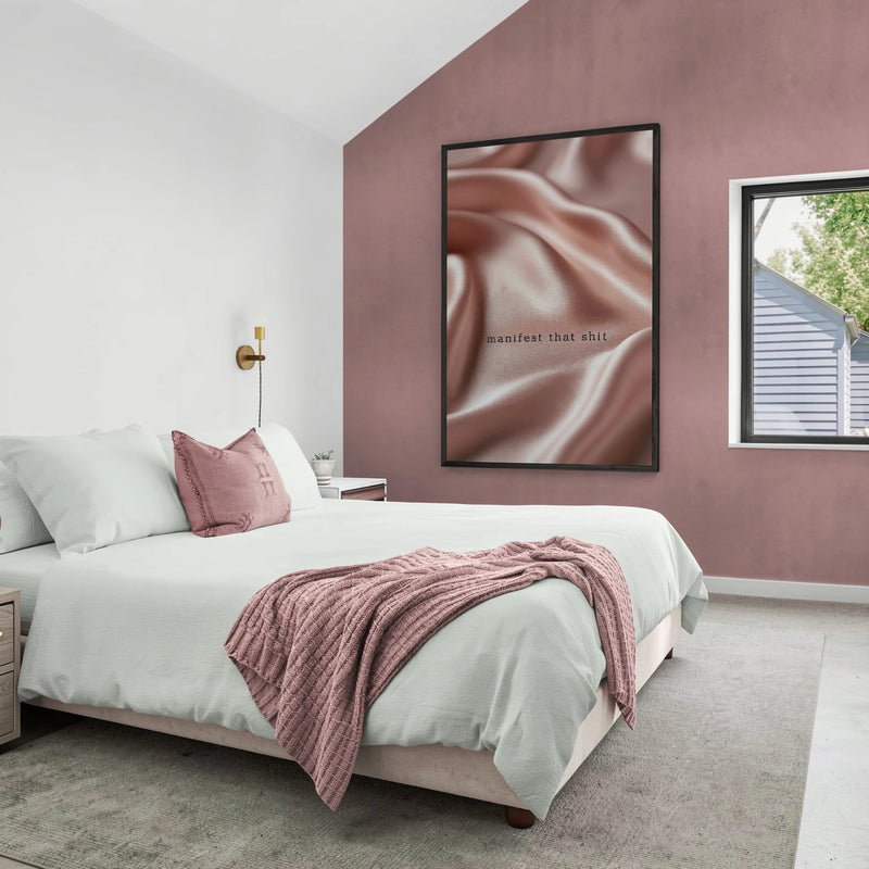 A framed wall art print featuring a close-up of silky satin fabric in dusty rose and brown tones, hanging on a mauve-pink wall in a modern bedroom setting. The phrase Manifest That Shit in tiny black text.