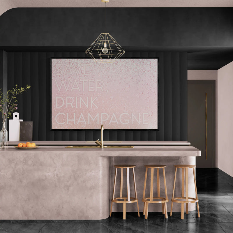 A modern kitchen interior with a large square wall art print featuring the words 'SAVE WATER DRINK CHAMPAGNE' in white text on a pale pink textured background. The print is mounted above a concrete-topped island with three wooden bar stools, set against a dark paneled wall.