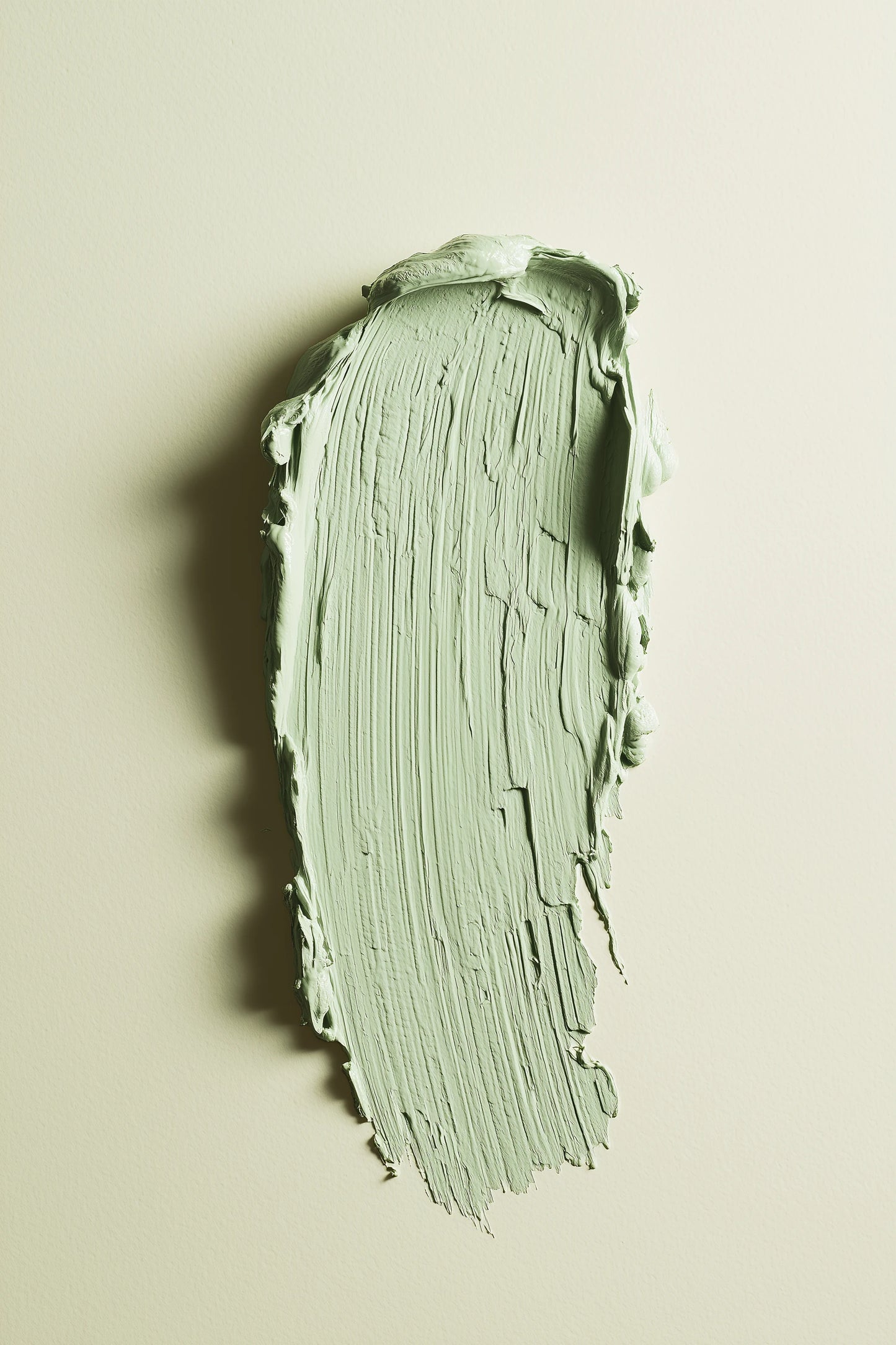 A textured swipe of pale sage green paint on a warm off-white background, showing the thick, creamy consistency and brushstroke texture of the product.