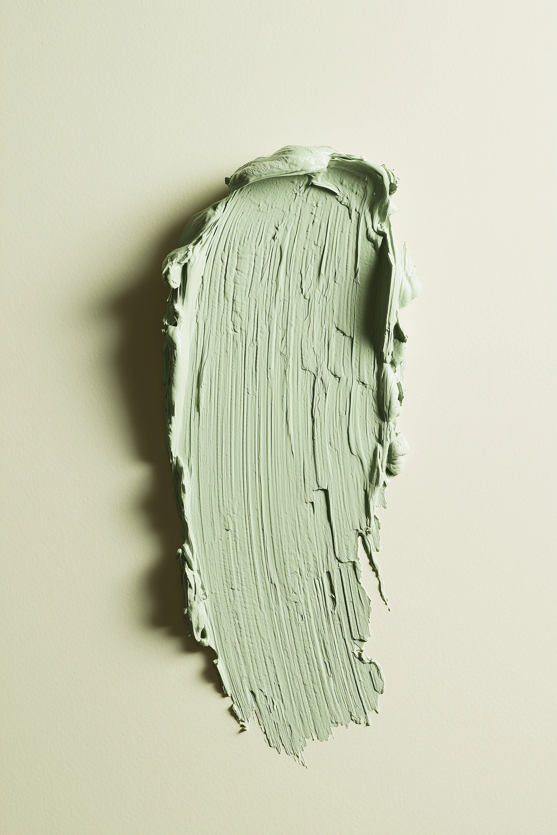 A textured swipe of pale sage green paint on a warm off-white background, showing the thick, creamy consistency and brushstroke texture of the product.