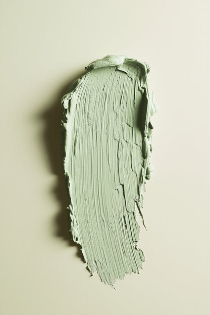 A textured swipe of pale sage green paint on a warm off-white background, showing the thick, creamy consistency and brushstroke texture of the product.