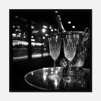 Black framed print of two wine glasses and a bottle of champagne in an ice bucket on a reflective surface with a night-time cityscape background.