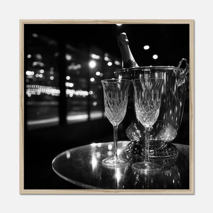 Natural oak framed print of two wine glasses and a bottle of champagne in an ice bucket on a reflective surface with a night-time cityscape background.