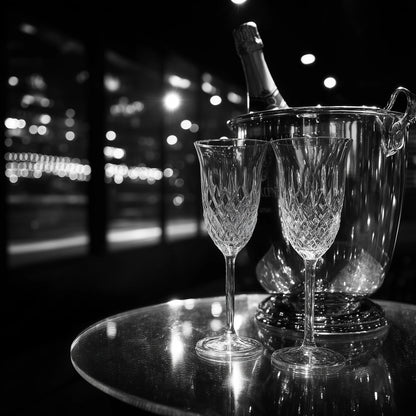 A black and white photograph of a crystal ice bucket with a champagne bottle inside and two cut-crystal champagne flutes on a reflective round table, set against a blurred nighttime city window background.