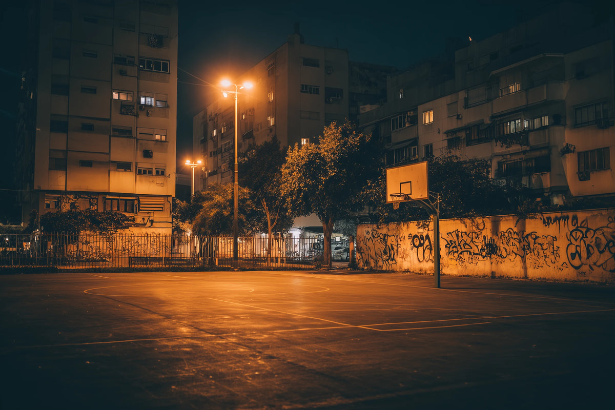 A moody night photograph of an empty outdoor basketball court in an urban setting, illuminated by warm yellow streetlights. Surrounding the court are graffiti-covered walls, trees, and multi-story residential buildings under a dark night sky.