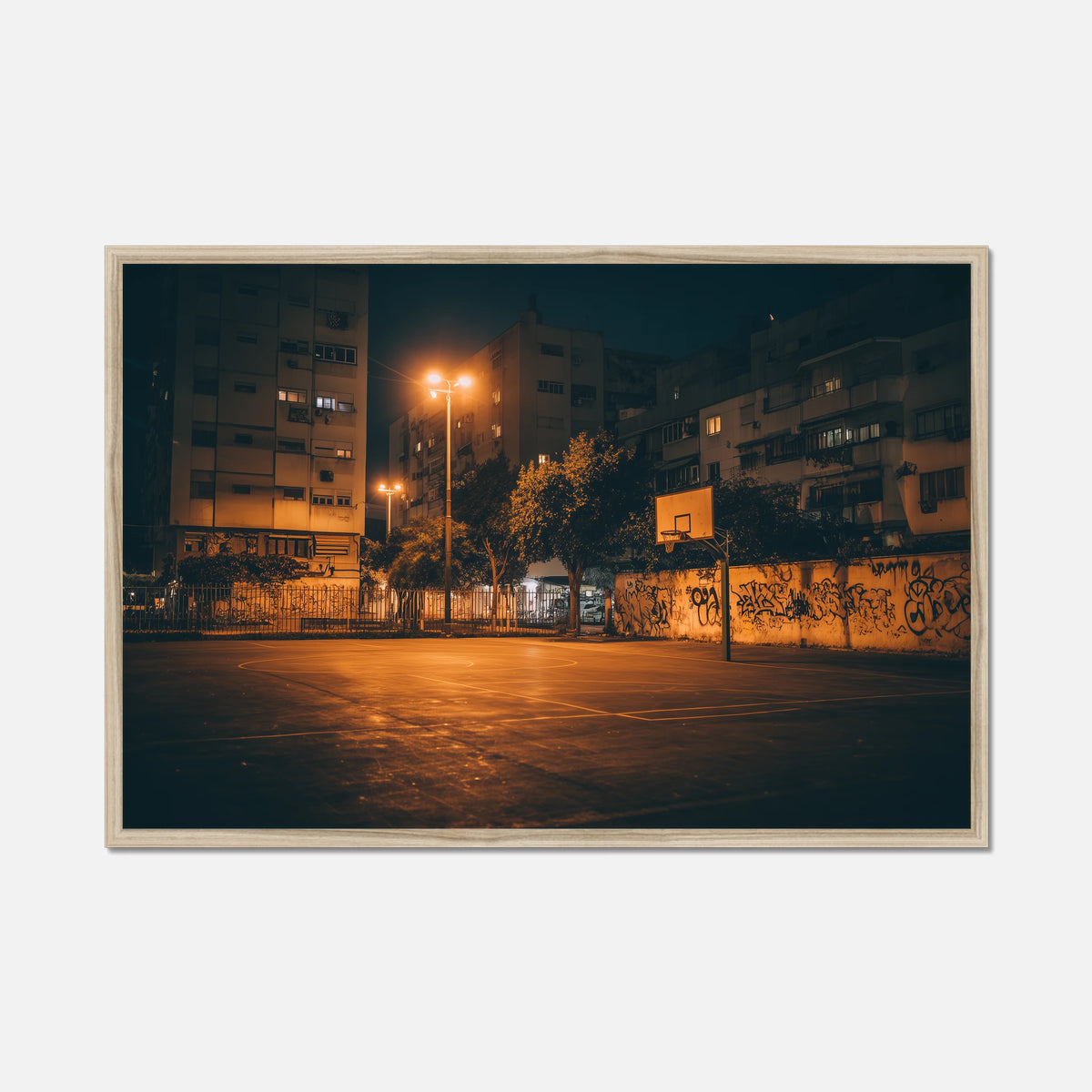A framed photographic print of an empty urban basketball court at night, illuminated by warm streetlights with apartment buildings and graffiti walls in the background, shown against a light gray backdrop.