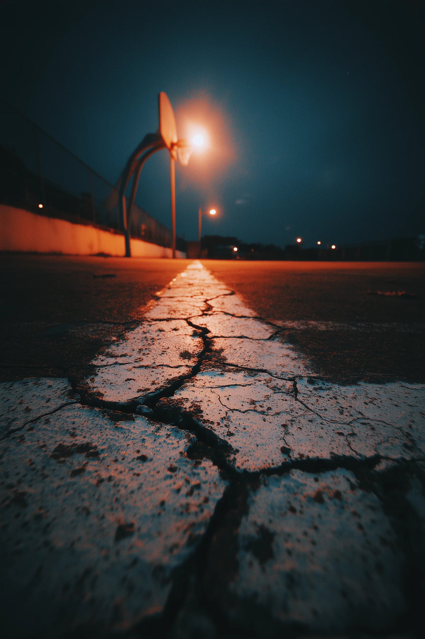 A low-angle night photograph of a cracked asphalt road with a faded white line, illuminated by a warm streetlight under a dark blue sky.