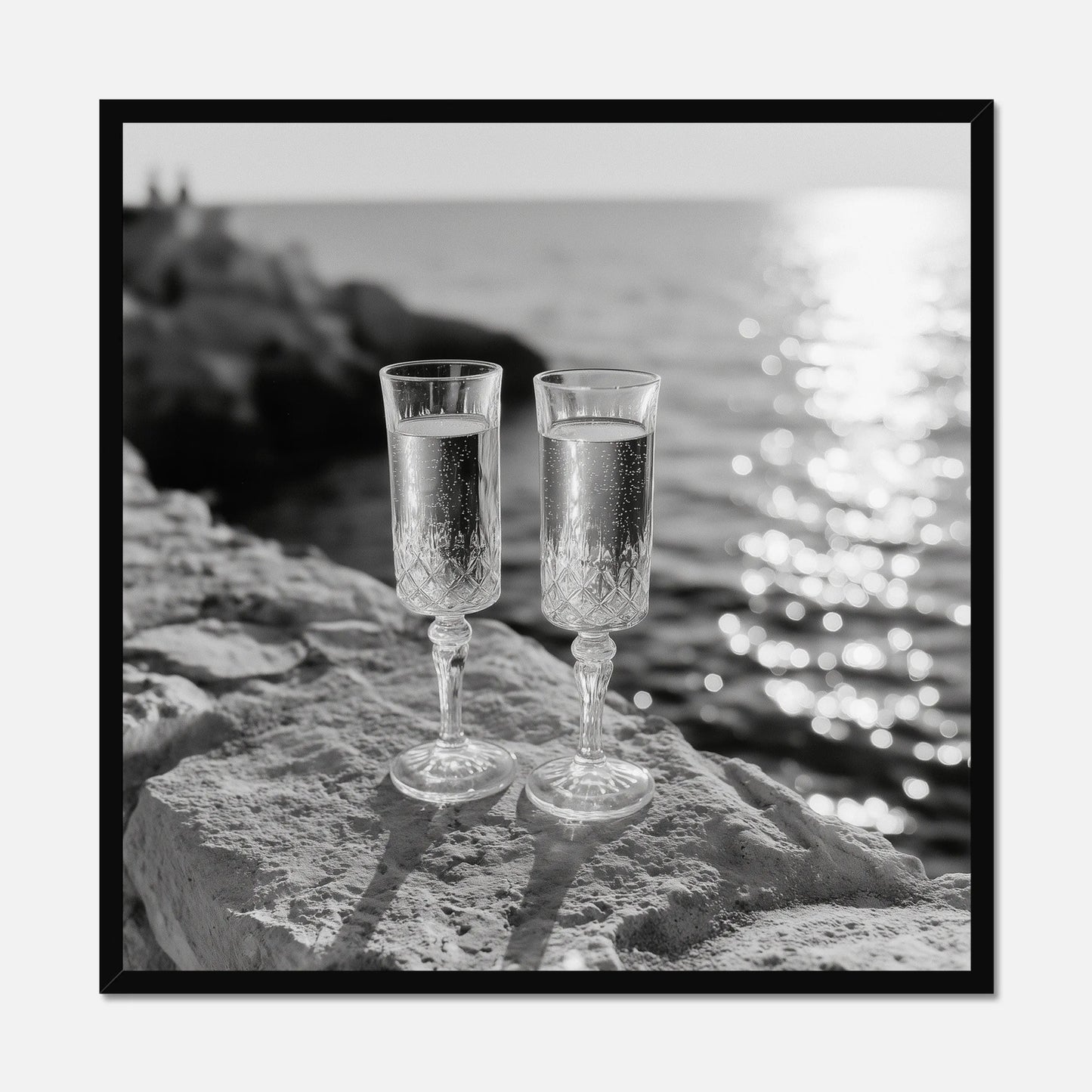 Two champagne glasses on a rock by the water displayed in a black wooden frame.