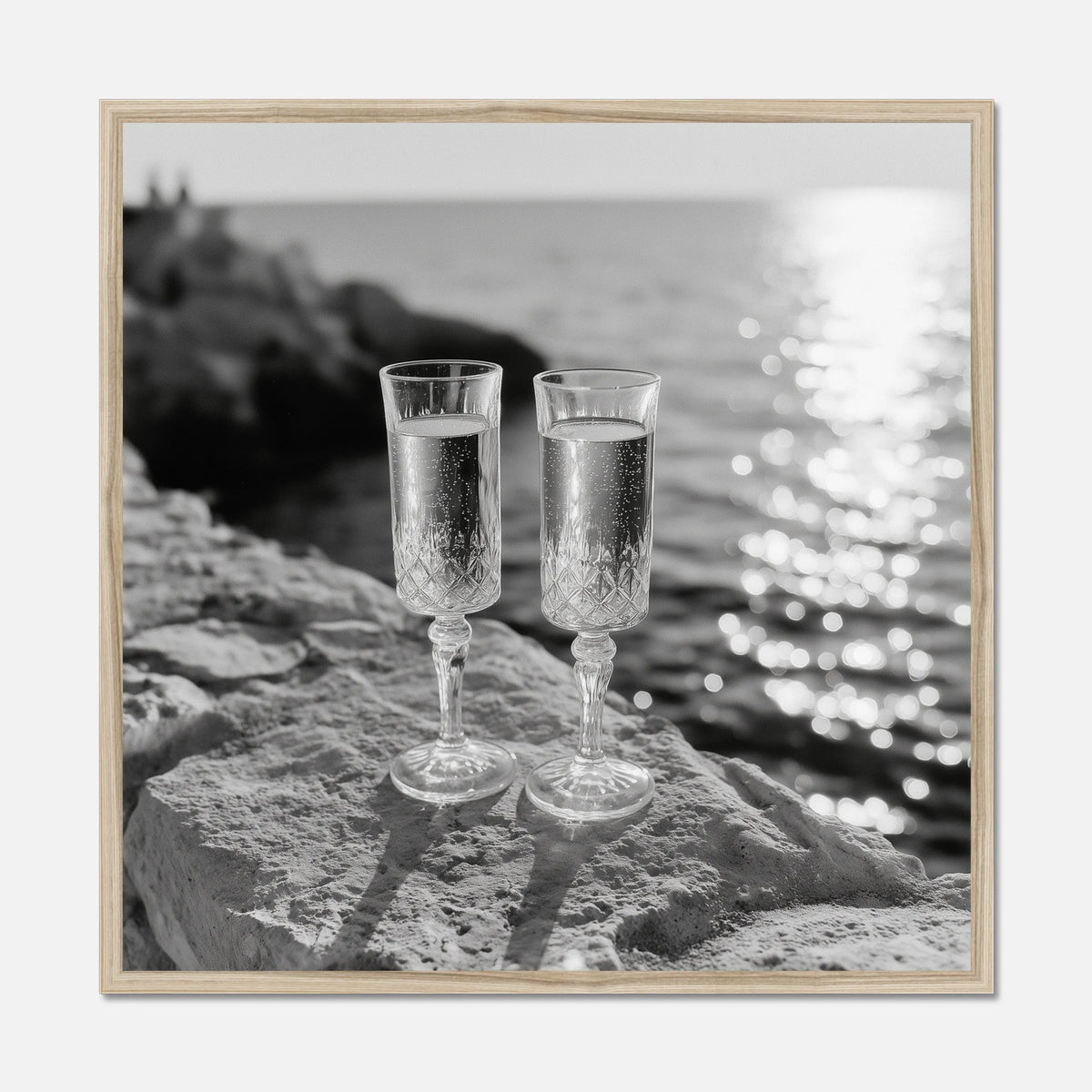 Two champagne glasses on a rock by the water displayed in a natural oak wooden frame.