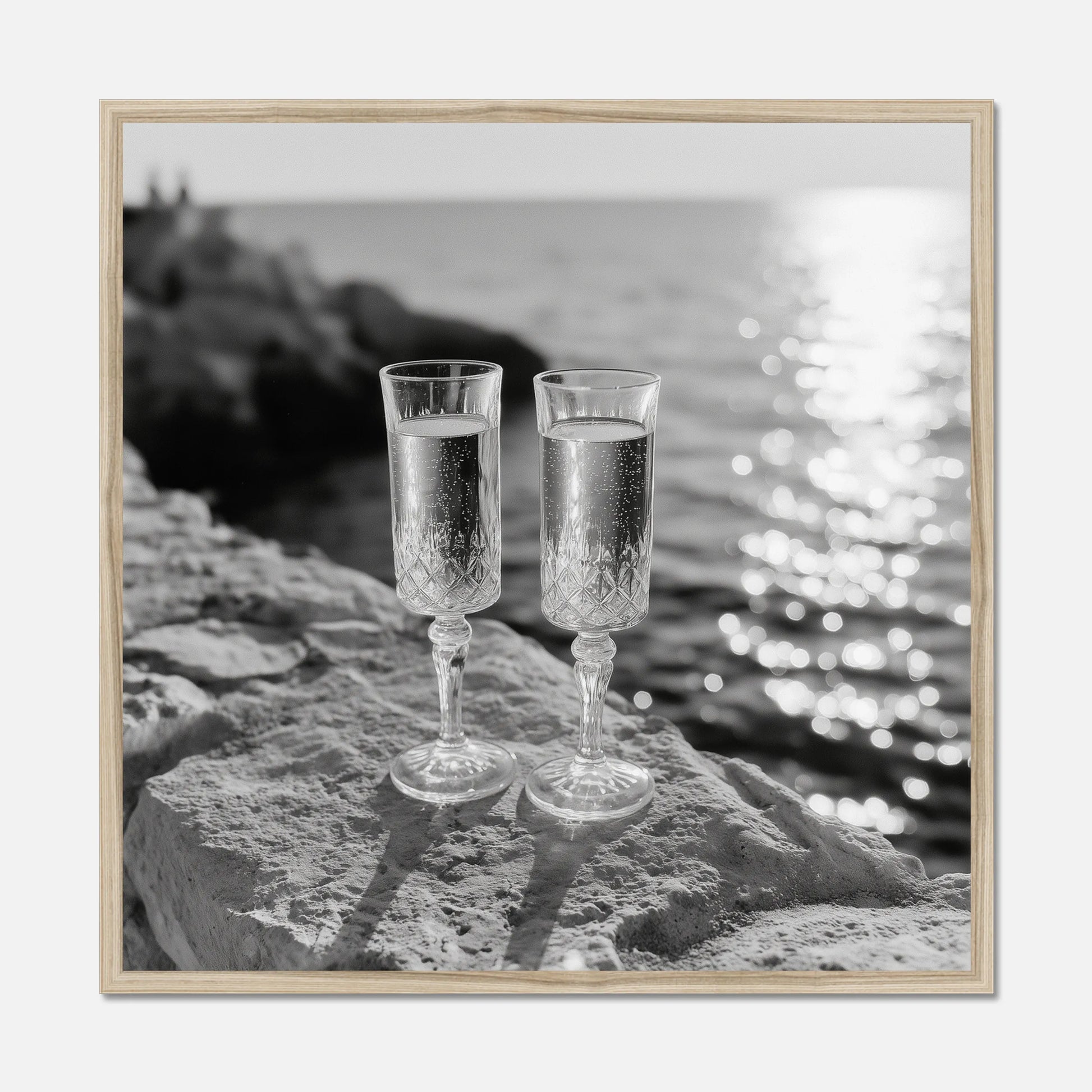 Two champagne glasses on a rock by the water displayed in a natural oak wooden frame.