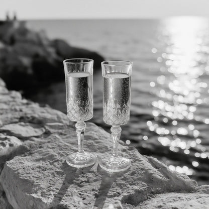 Two clear champagne flutes filled with a bubbly drink, set on a stone ledge with a blurred ocean and rocky shoreline in the background.