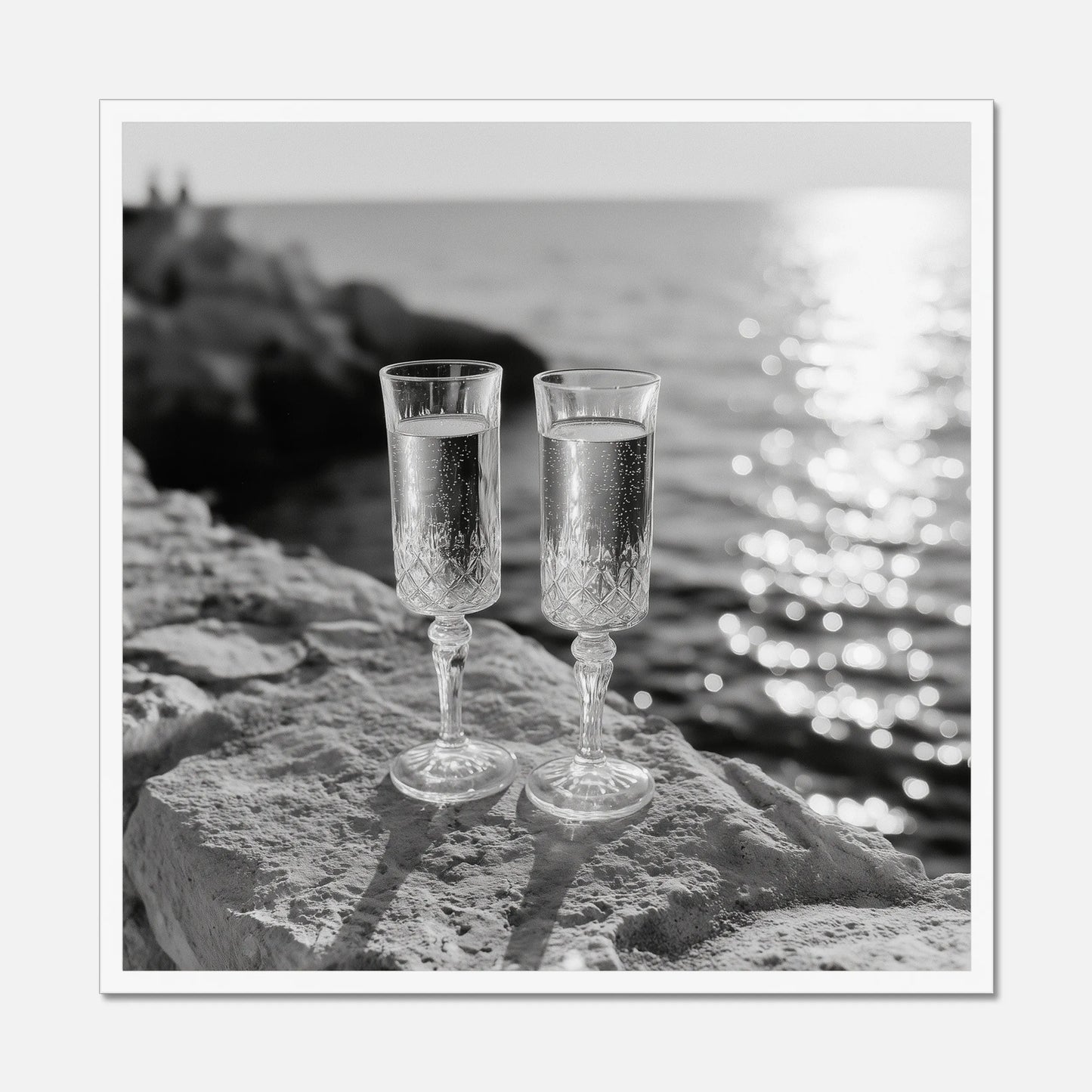 Two champagne glasses on a rock by the water displayed in a white wooden frame.