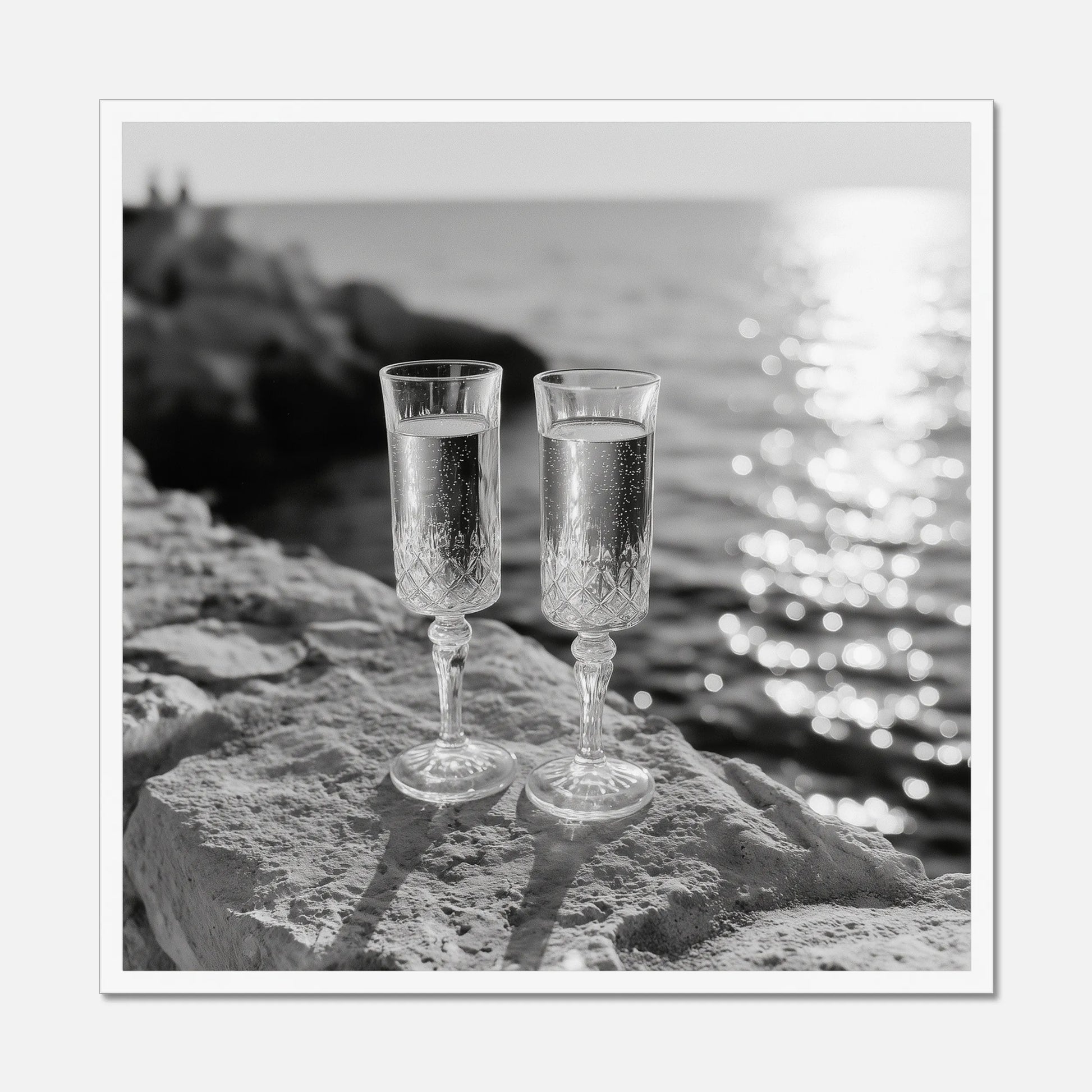 Two champagne glasses on a rock by the water displayed in a white wooden frame.