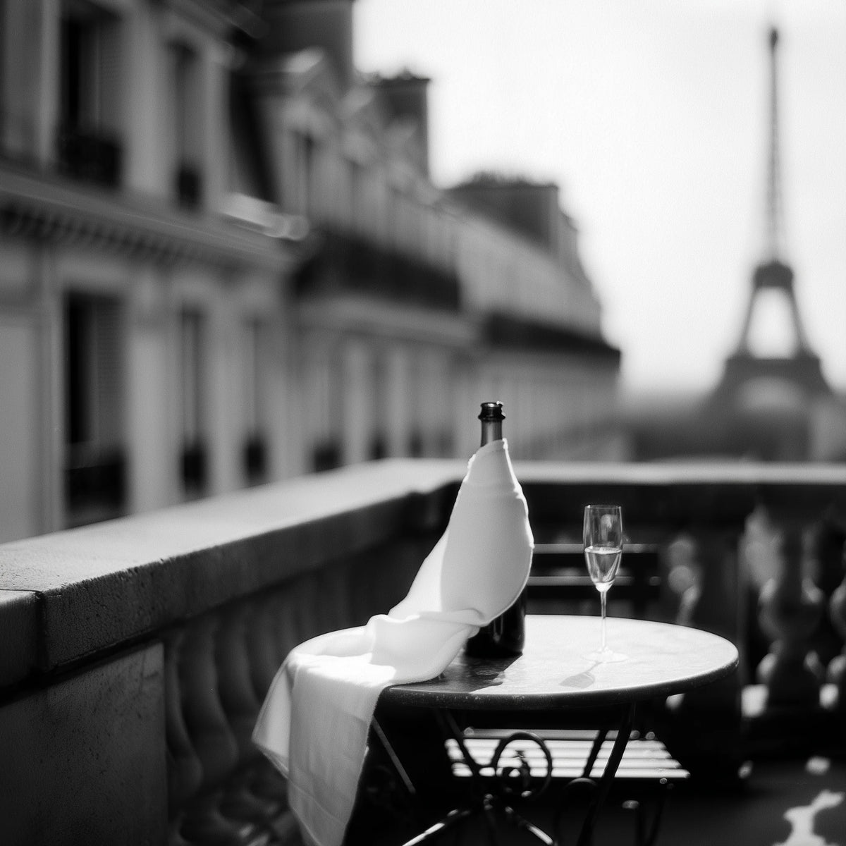 A black and white photograph of a Parisian balcony scene with a bottle and a folded cloth on a small round table, with a wine glass and a blurred Eiffel Tower in the background.