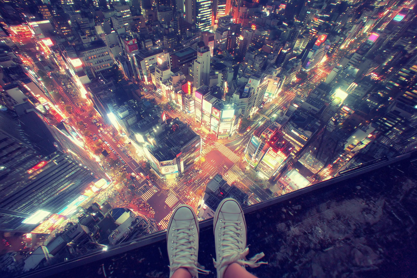 A high-angle night photograph of a crowded Tokyo city skyline with glowing streetlights and buildings, viewed from a high vantage point with a person's feet in white sneakers resting on the edge.