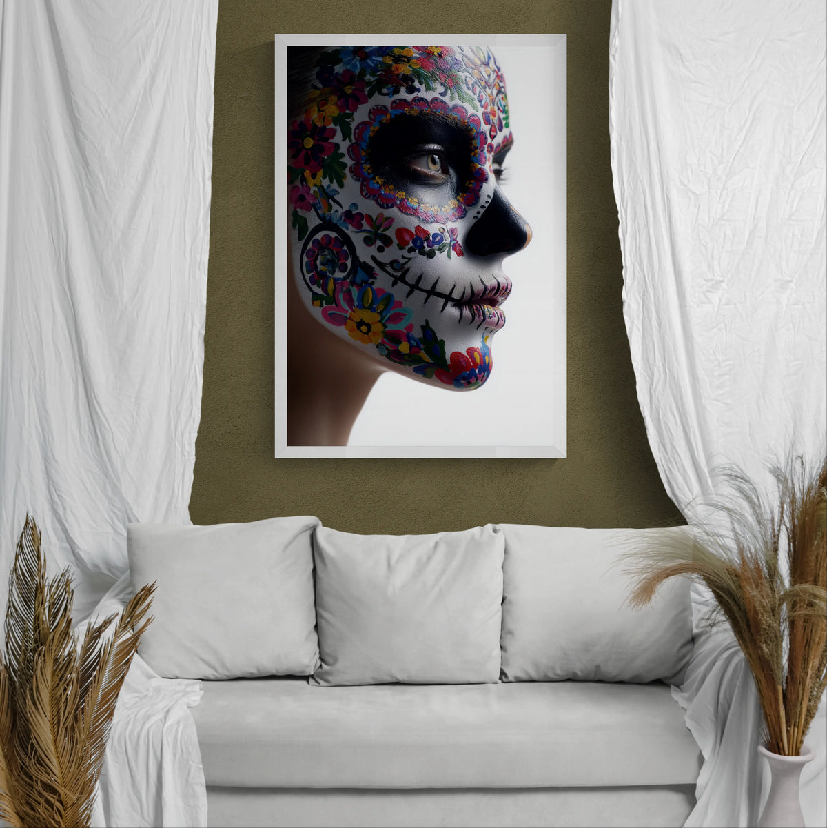 A framed wall art print showing a woman's face in colorful floral sugar skull makeup, displayed above a white sofa in a styled room with white drapes and pampas grass.