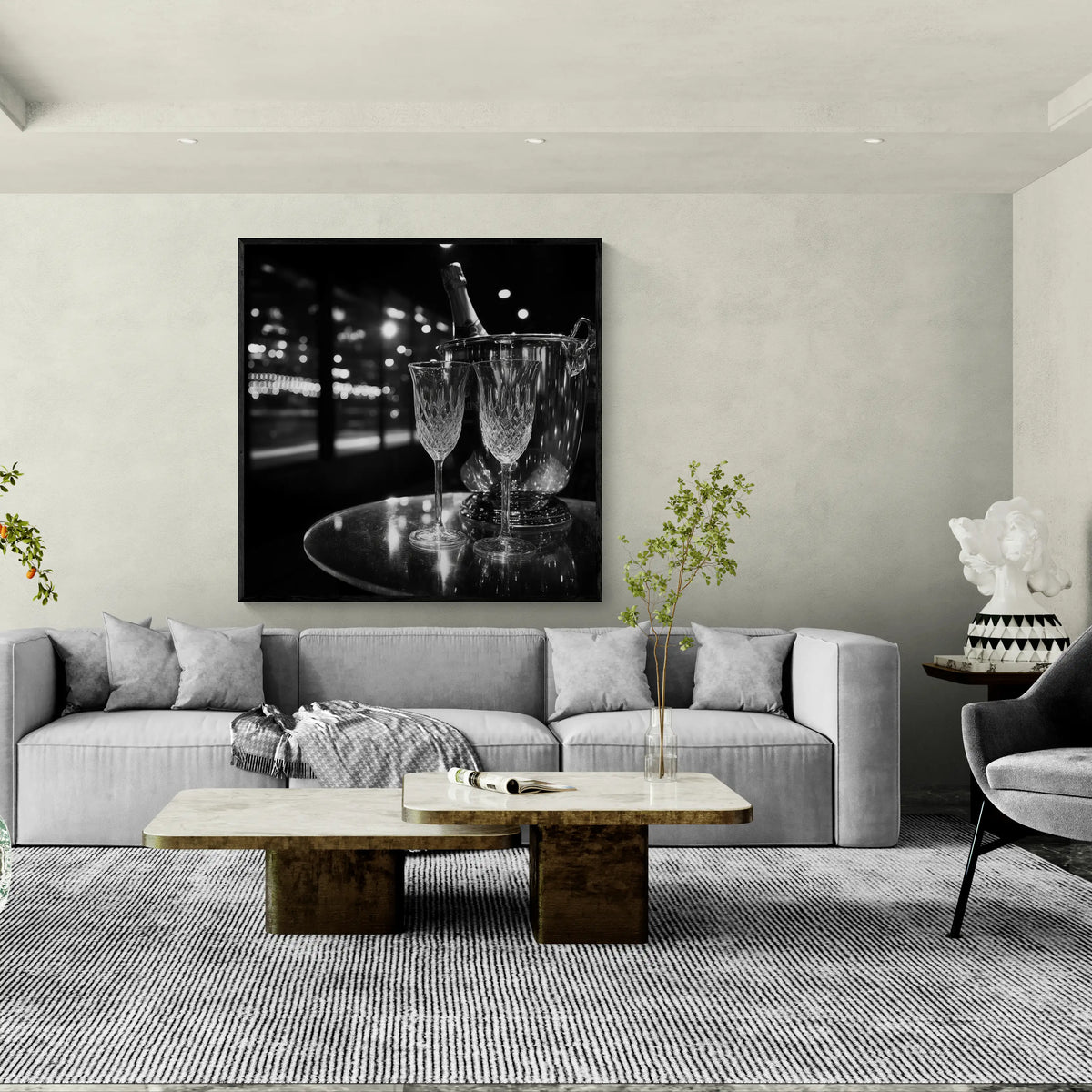 A large black and white wall art print depicting a champagne and wine glass scene, displayed above a modern gray sofa in a styled living room setting.
