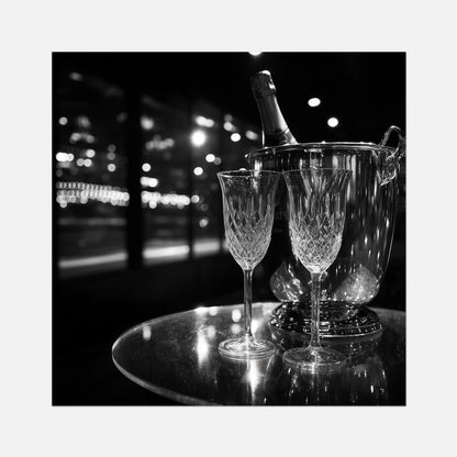 Fine art print of two wine glasses and a bottle of champagne in an ice bucket on a reflective surface with a night-time cityscape background.