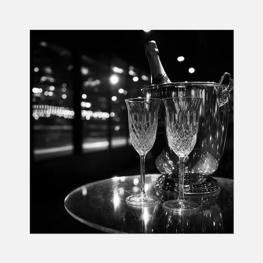 Fine art print of two wine glasses and a bottle of champagne in an ice bucket on a reflective surface with a night-time cityscape background.