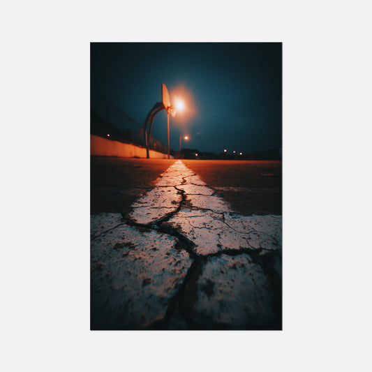 A vertical photographic art print depicting a cracked asphalt road at night with a bright streetlight casting a warm orange glow, centered on a light gray background.