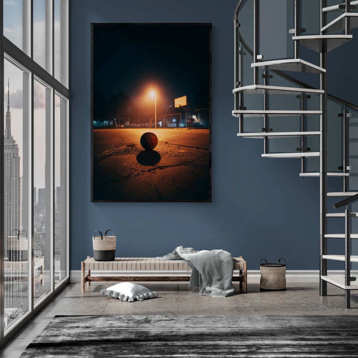 A large vertical framed wall art print depicting a moody nighttime basketball court scene with a glowing streetlamp and shadow, displayed on a blue-gray wall in a modern interior with a spiral staircase, bench, and city view.