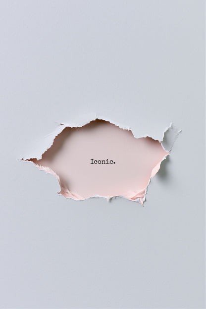 A minimalist art print featuring a torn paper hole in the center of a light gray background, revealing a pale pink paper layer underneath with the word 'Iconic.' printed in black typewriter font.