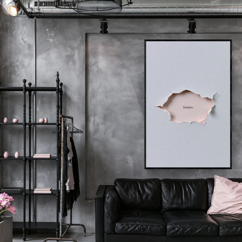 Typography artwork in a industrial lounge. A framed wall art print hangs on a gray concrete wall above a black leather sofa in a modern living room. The print shows a light gray background with a torn-paper effect revealing a pale blush pink interior and the word 'Iconic' in small script.