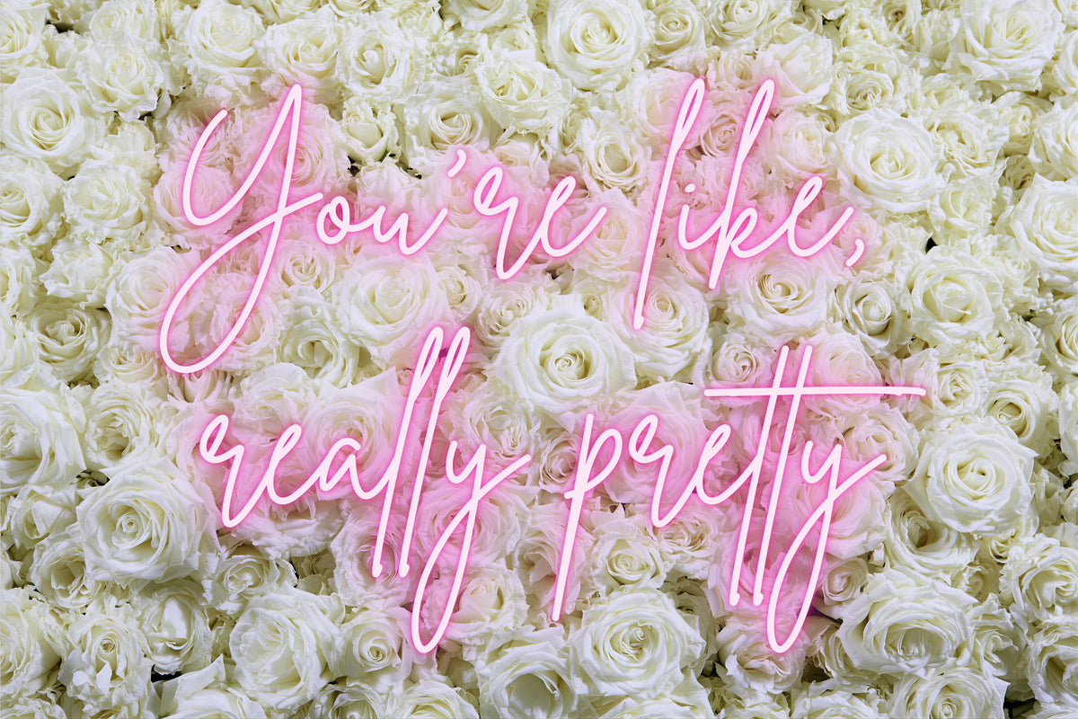 A dense wall of white roses with pink neon-style script text reading 'You're like, really pretty' across the center.