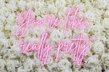 A dense wall of white roses with pink neon-style script text reading 'You're like, really pretty' across the center.