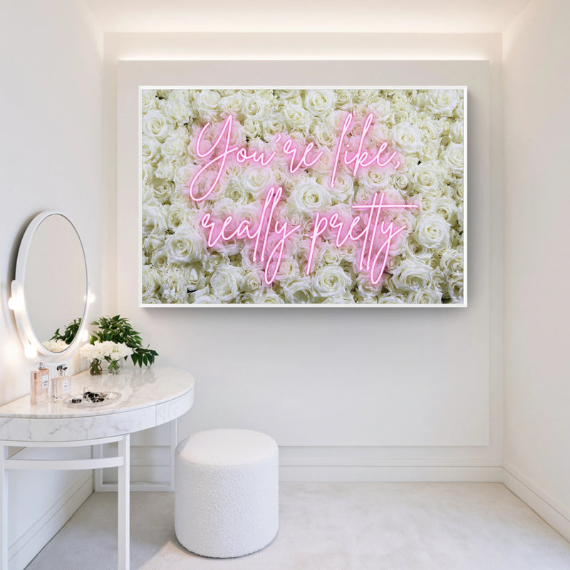 A framed wall art print featuring a white rose background with pink neon-style script text reading 'You're like, really pretty,' displayed in a bright, modern vanity room setting.